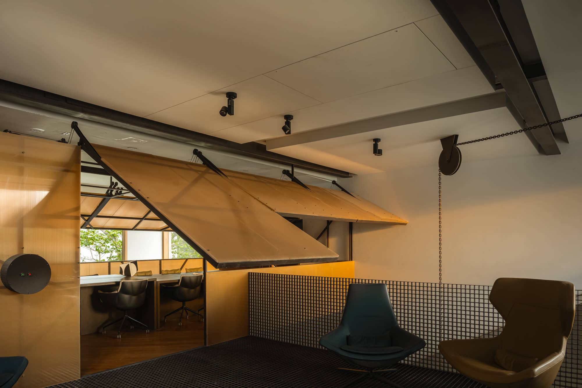 Folded plywood canopy panels suspended over a workspace, with lounge chairs in the foreground — daylight