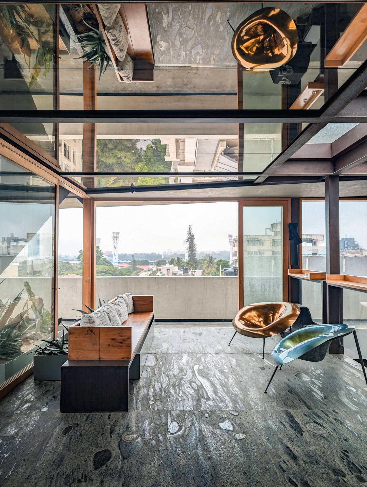 Glass-walled terrace corner with brass pendants, low seating, and the city skyline beyond — daylight