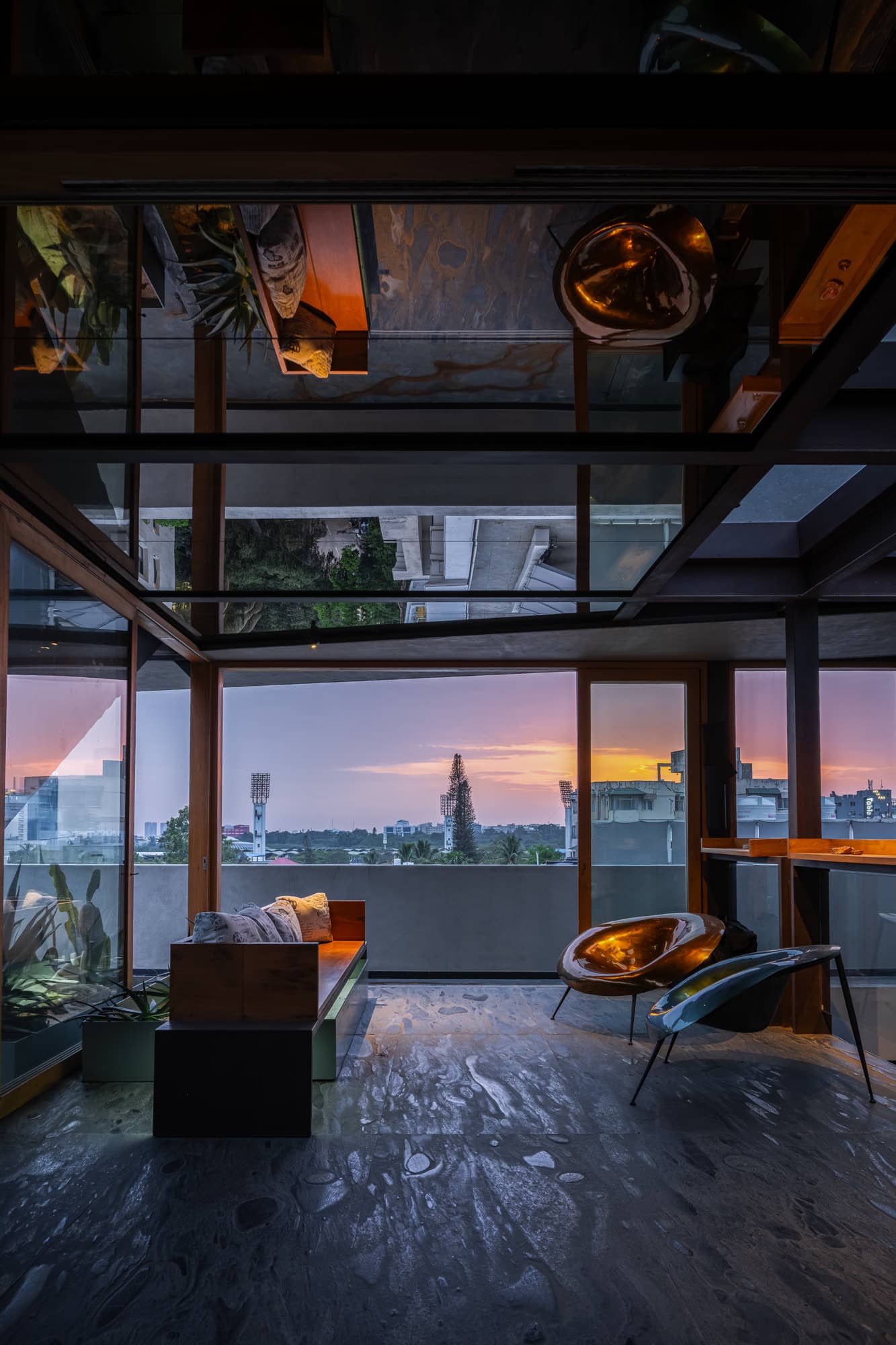 Glass-walled terrace corner with brass pendants, low seating, and the city skyline beyond — after dark